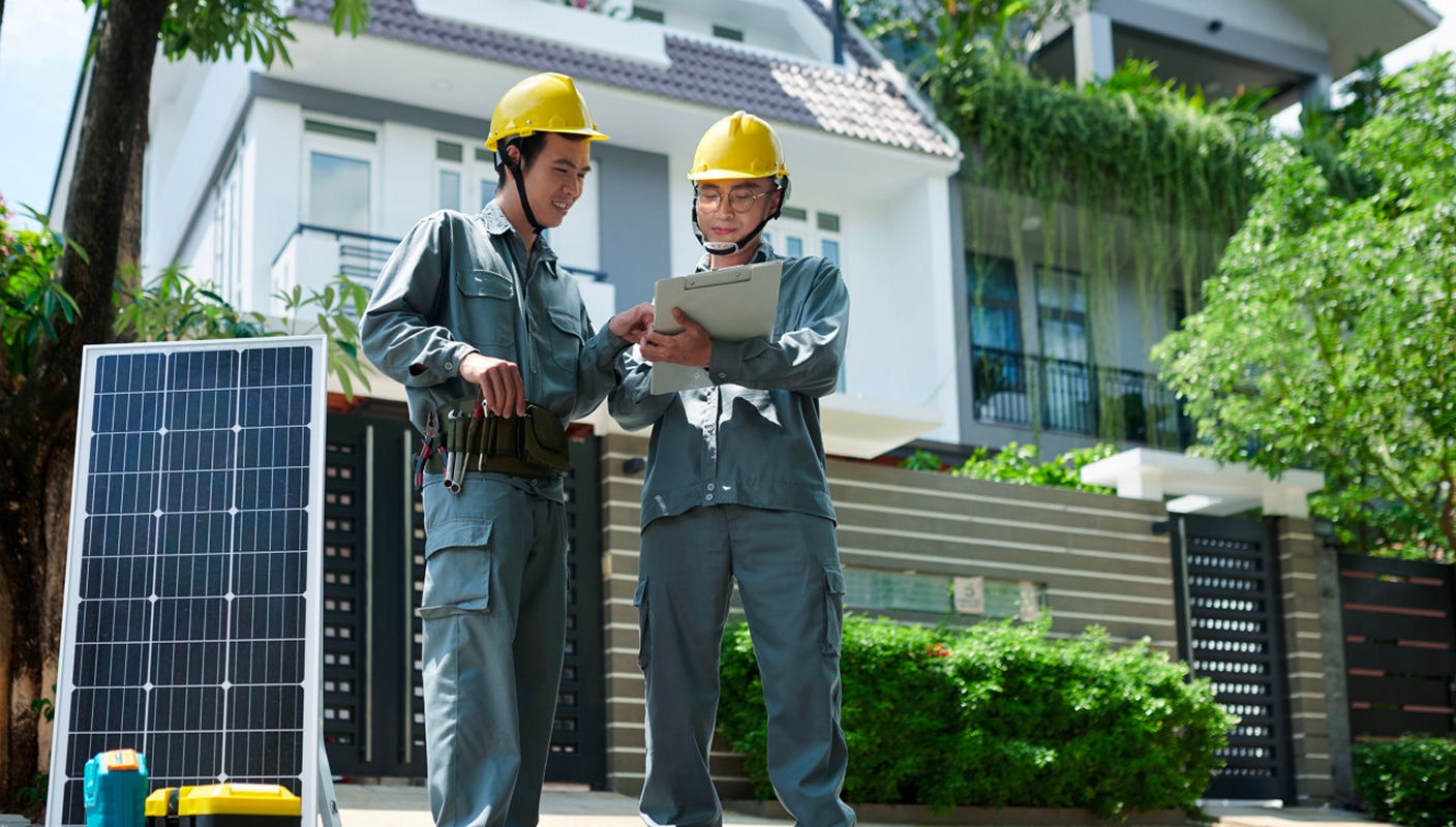 Two engineers with safety helmets discussing solar project progress on site with solar panels in background, holding tablet and clipboard