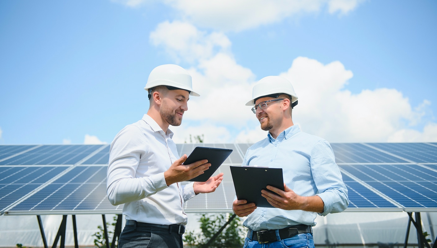 Professionals reviewing solar energy project details with tablet and clipboard near solar panel installation under a clear sky