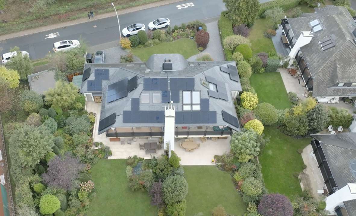 Aerial view of a premium residential home with an integrated solar rooftop system, utilizing full-black PV panels across a multi-angled roof design, surrounded by a landscaped garden