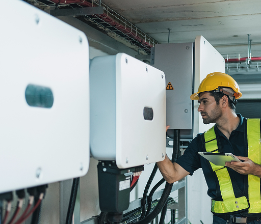 A male technician wearing a yellow hard hat and reflective vest is inspecting solar inverters mounted on a wall while holding a tablet. He is working in an indoor technical facility with cables and electrical equipment.