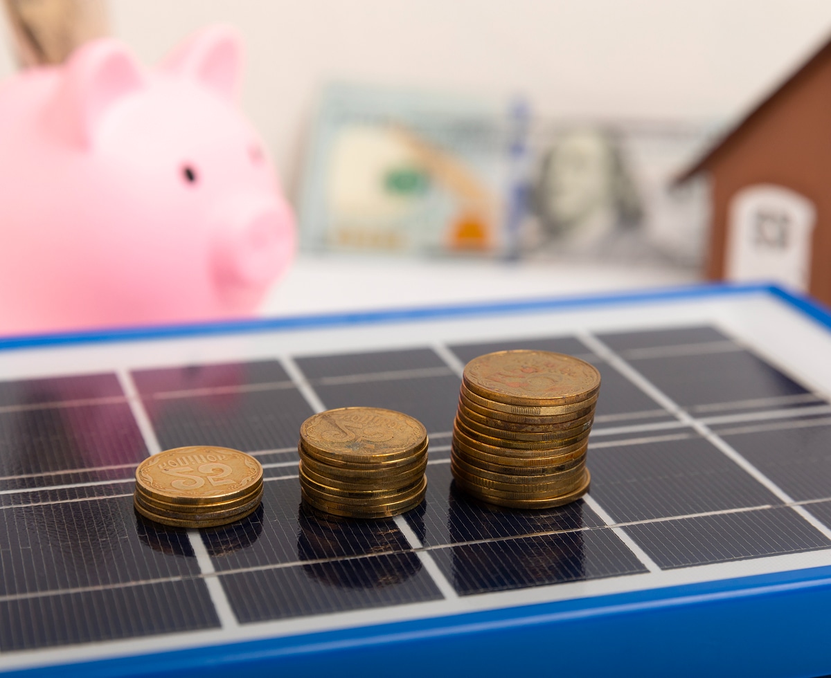 Stacks of coins on a miniature solar panel with piggy bank and house model in the background, symbolizing solar investment and savings
