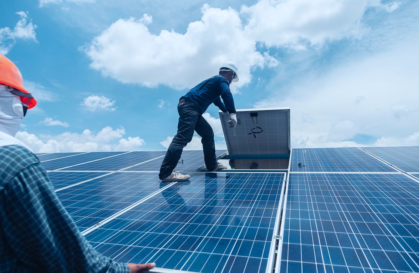 Solavita Solar panel installation workers on the roof, one lifting a panel while the other assists, with a clear sky in the background.